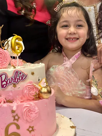 The birthday girl smiling with her two-tier Barbie cake. The pink rosettes and gold details make it a truly glamorous centerpiece.