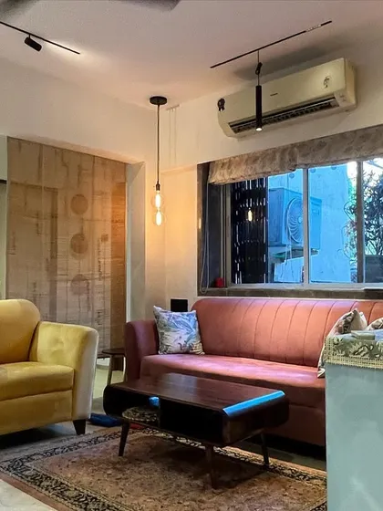 An eclectic living room with a mix of furniture styles, including a pink mid-century modern sofa and a yellow armchair. The lighting combines track lights with a decorative pendant light.