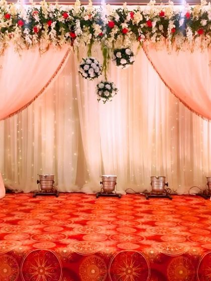 A classic stage backdrop with peach-colored drapes, a canopy of white and red-tinged flowers, and hanging floral spheres, creating a timeless and romantic setting.