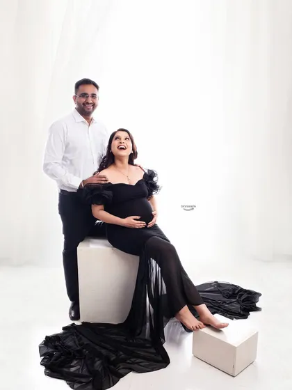 A joyful and relaxed couple portrait. The mom-to-be is seated in a beautiful black gown while her partner stands behind her, sharing a happy moment in a bright, high-key setting.