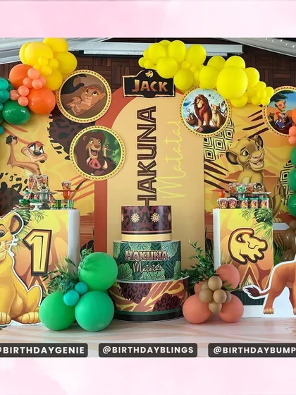 A vibrant "Hakuna Matata" first birthday setup, featuring characters from The Lion King. The decor includes multiple themed plinths for the cake and desserts, character cutouts, and a colorful balloon garland.