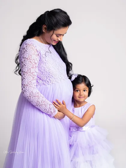 A mother and daughter duo, beautifully coordinated in matching lavender gowns. The older sister-to-be is so excited, gently holding her mother's belly in this sweet studio maternity portrait.