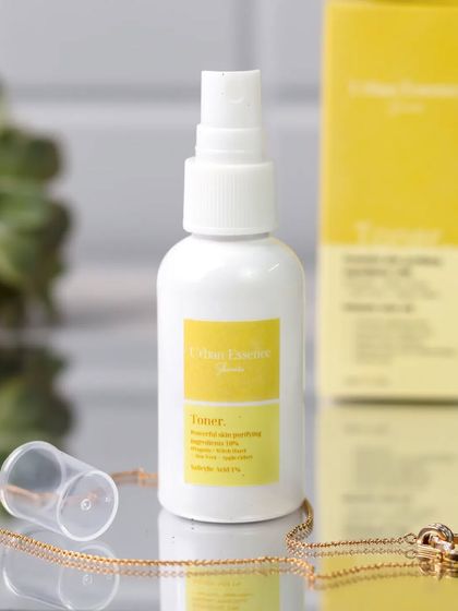 A toner bottle is styled with a simple gold necklace against a bathroom tile background. The yellow of the label and the gold accessory create a cohesive and bright color story.