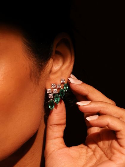 A model wearing the Emerald Cascade earrings, showing how they elegantly frame the face. The combination of princess-cut diamonds and pear-shaped emeralds creates a balanced, brilliant design.