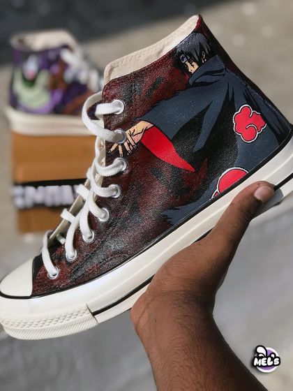 A hand-held shot of the Itachi Uchiha custom Converse. The design captures his iconic Akatsuki cloak and pose against a textured, dark red and black background.