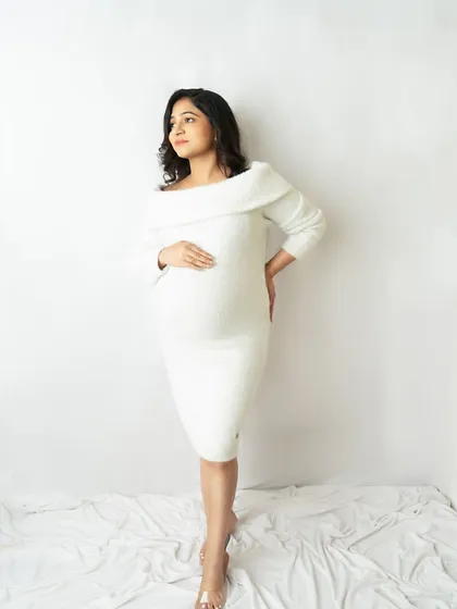 A full-length portrait in a chic, off-the-shoulder white sweater dress. The clean, minimalist background keeps the focus on you and your bump.