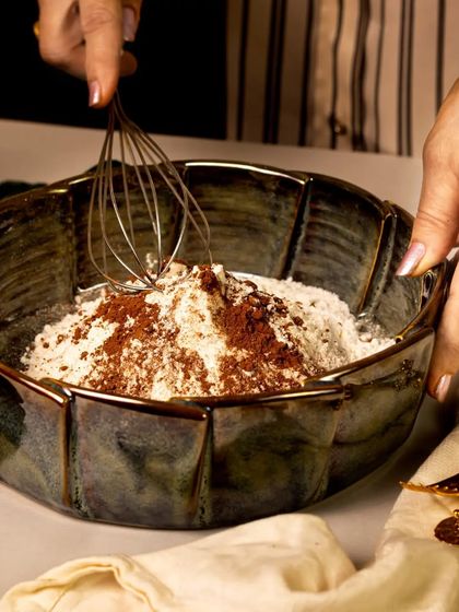 This lifestyle shot captures the joy of baking, with hands whisking flour and cocoa in a beautiful rustic ceramic bowl. The image feels warm, authentic, and full of creative energy.