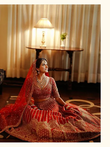 A beautifully lit indoor portrait of a bride sitting gracefully, her red lehenga spread around her, creating a warm and regal image.