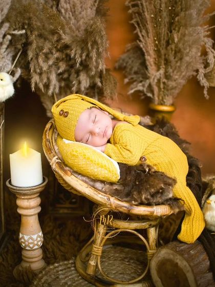 A newborn in a yellow knit outfit sleeps on a miniature chair in a rustic, warm-toned studio setup.