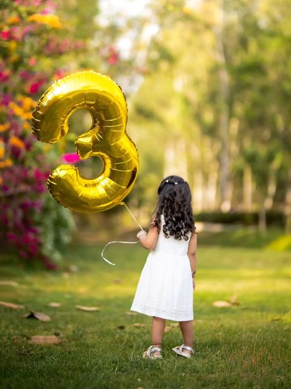 A creative shot for a third birthday. This outdoor portrait captures a little girl with a '3' balloon, marking her special day in a beautiful park setting.