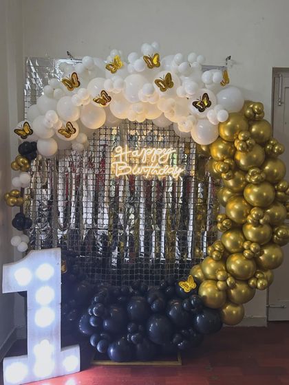 An elegant and chic first birthday setup. The circular balloon arch in black, gold, and white is accented with butterfly details and a shimmering silver backdrop. The light-up number '1' adds a touch of glamour to the celebration.