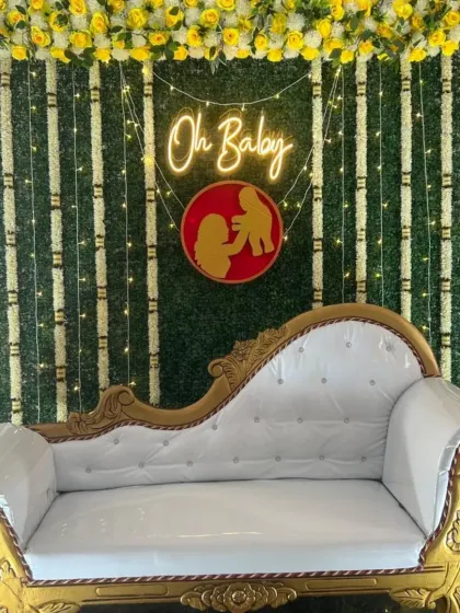 An elegant baby shower stage with a traditional touch. The setup features a beautiful white and gold chaise lounge set against a green foliage wall with an "Oh Baby" neon sign and floral garlands.