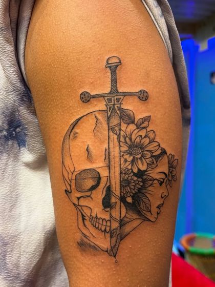 This tattoo blends life and death, with a skull on one side and a woman's face with flowers on the other, divided by a sword. A powerful piece about duality.