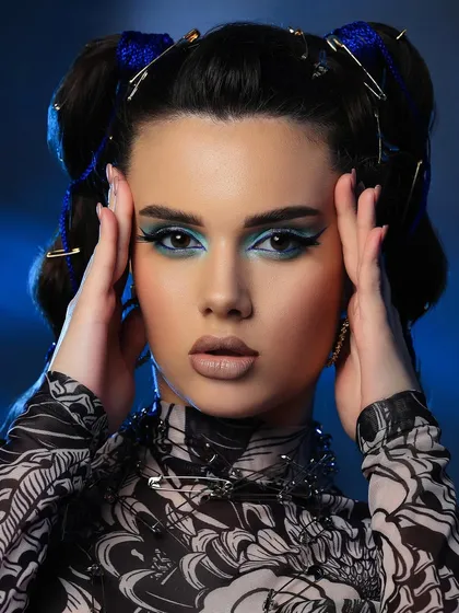 A creative beauty portrait with a model wearing a safety pin necklace and lip cuff, with bold blue eye makeup against a blue-lit background.