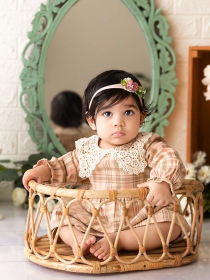 A baby girl sits in a wicker basket in a vintage-themed setup with a mirror and floral accents.