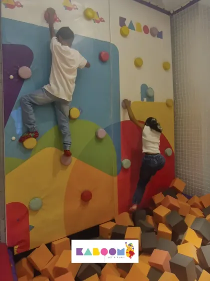 Two friends tackle the climbing wall together, with a soft foam pit waiting below.