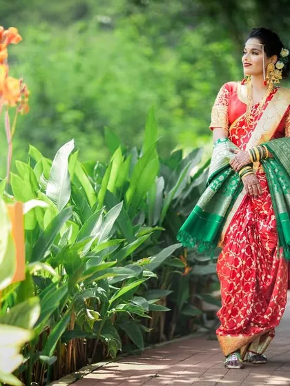 The bride walks through a garden path, her vibrant red and green saree standing out against the lush foliage.