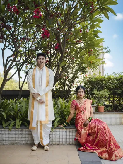 A beautifully composed portrait of Roshan and Meenakshi. The bride is seated while the groom stands, creating a classic and elegant composition that showcases their wedding outfits and the lovely outdoor setting.