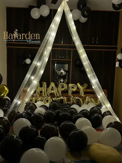 Creating a romantic and cozy birthday surprise right in the bedroom. I use a canopy made of sheer drapes and fairy lights over the bed, surrounded by dozens of black and white balloons. The 'Happy Birthday' foil balloons complete this intimate celebration.