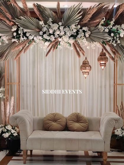 A bohemian-inspired reception stage featuring dried palm leaves, pampas grass, and white florals, creating a textured and earthy backdrop with hanging lanterns for a warm glow.