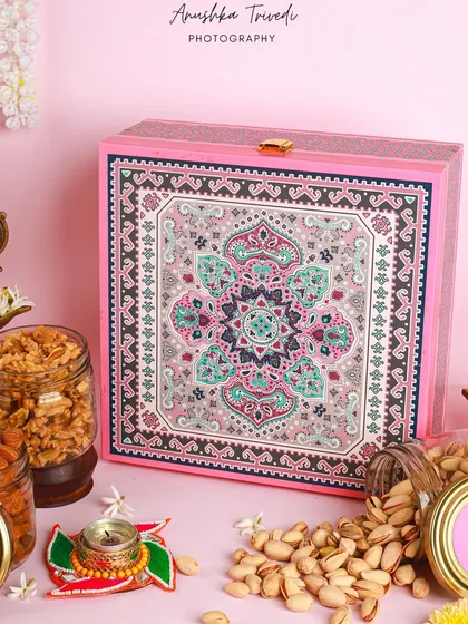A stunning pink and teal Diwali hamper filled with jars of nuts and snacks. The styling with brass lamps and garlands of jasmine adds a touch of traditional elegance.