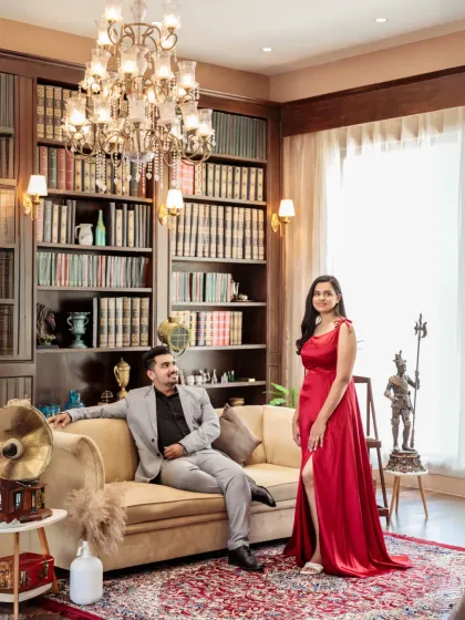 A sophisticated portrait in a classic library setting. The couple exudes elegance, with the bride in a striking red gown and the groom looking dapper, surrounded by books and a crystal chandelier.