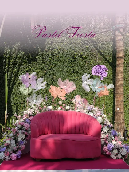 A 'Pastel Fiesta' photo booth. The vibrant pink sofa is set against a wall of green foliage, adorned with large, dreamy paper flowers in pastel shades, creating a perfect spot for Instagrammable photos.