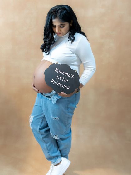A sweet and personal studio shot. The mom-to-be is wearing a casual outfit and holding a "Mumma's little Princess" sign on her baby bump.