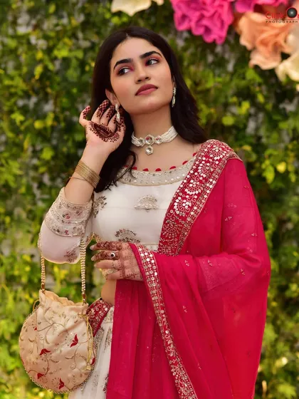 A fashion-forward portrait for a pre-wedding event. The model's pose highlights her elegant white and red lehenga and matching potli bag against a vibrant floral backdrop.