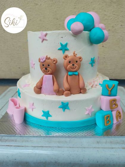 A sweet two-tier cake perfect for a twin celebration or baby shower. It features cute fondant teddy bears, stars, and baby blocks in a classic pink and blue color scheme.