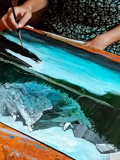 A close-up of a student painting a landscape, demonstrating the technique for creating realistic water reflections.