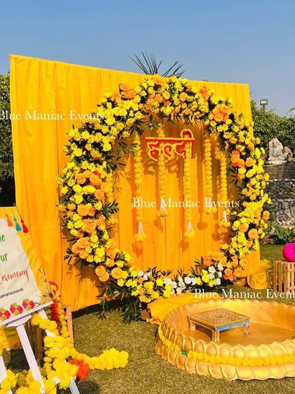 An outdoor Haldi ceremony setup featuring a circular floral wreath of marigolds against a bright yellow backdrop. The arrangement includes a personalized welcome sign and traditional seating for the rituals.