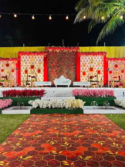 A grand and opulent reception stage with a red and gold theme. The backdrop features intricate patterns and a wall of red roses, creating a truly royal setting for the couple.