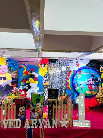A full stage setup for a Mickey Mouse first birthday. It includes a shimmer wall, rainbow accents, personalized backdrops, and large 'ONE' letters.