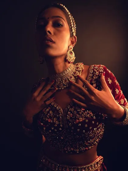 An artistic, low-light shot from a fashion photoshoot. The makeup focuses on creating a "glow from within" effect on the skin, complementing the rich velvet lehenga.