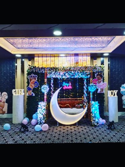 A baby shower stage in a hall with elegant, dark patterned walls. The setup includes a swing, moon prop, and 'Boy'/'Girl' signs.