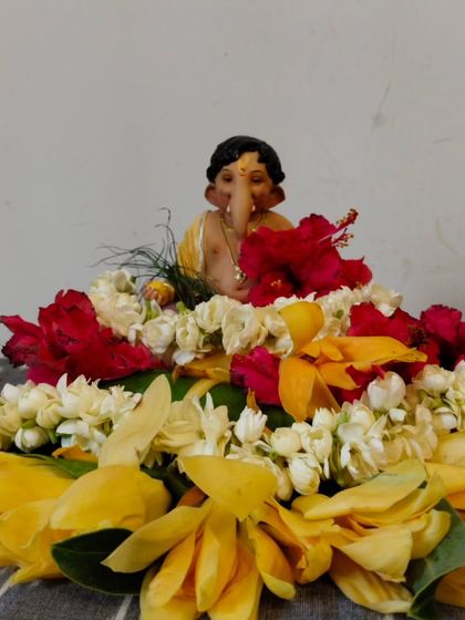 A small, adorable Ganpati idol seated on a bed of vibrant flowers, including hibiscus and jasmine. This simple and sweet arrangement is perfect for a personal desk or a small home altar.