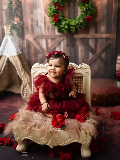 A happy baby girl in a rich, red dress, surrounded by rustic details and flowers. This milestone session has a warm and festive feel.