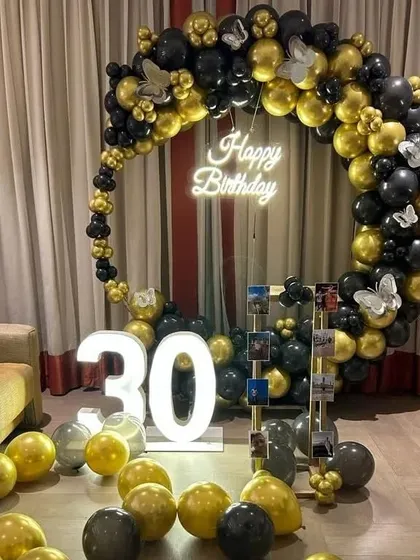 A 30th birthday party with a circular balloon arch in black and gold. The decoration includes a "Happy Birthday" neon sign, marquee numbers, and a photo display stand.