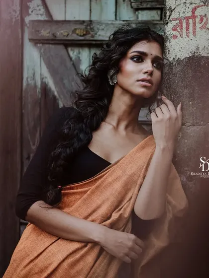 A moody, vintage-toned portrait of a model against a weathered wooden door. The textures of the wall and the soft drape of her orange saree create a rustic and soulful image.