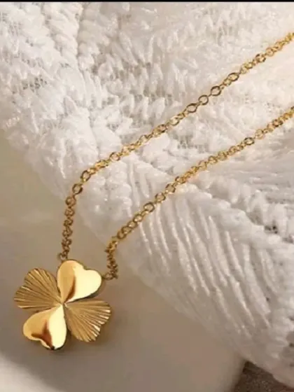 The gold-toned four-leaf clover necklace resting on a white lace fabric, showing its delicate chain and polished pendant. A beautiful gift for only 10 shekels.