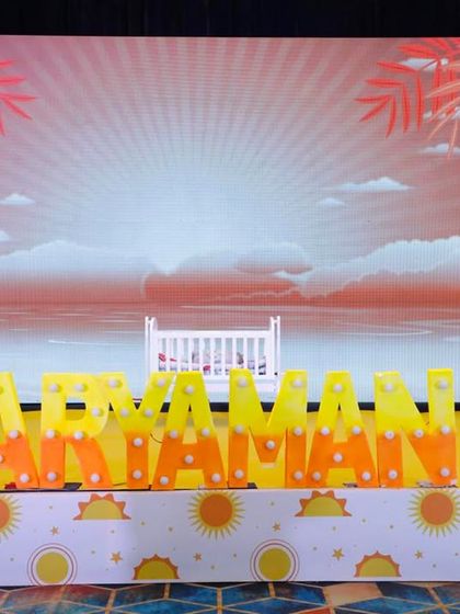 A closer look at the stage for Aryaman's naming ceremony. The backdrop features a serene beach sunrise, a tiny cradle, and the baby's name in bright marquee letters, creating a beautiful and meaningful scene.