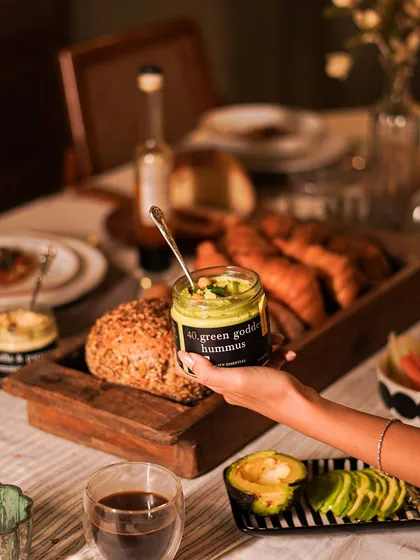 A jar of our vibrant Green Goddess Hummus, held over a beautiful breakfast spread. This creamy, herb-infused hummus is a healthy and delicious addition to any meal, perfect with fresh bread or vegetable sticks.