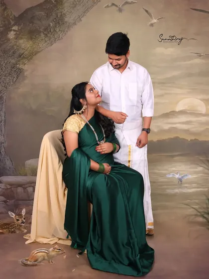A classic portrait of an expecting couple in traditional South Indian attire. The elegant green saree and white veshti create a beautiful contrast against the painterly backdrop.