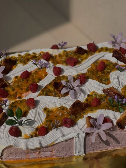 A close-up of our Passion Fruit Raspberry sheet cake. It has a vanilla sponge soaked in passion fruit juice, raspberry mousse, and is topped with fresh passion fruit, raspberries, and caramelized white chocolate.