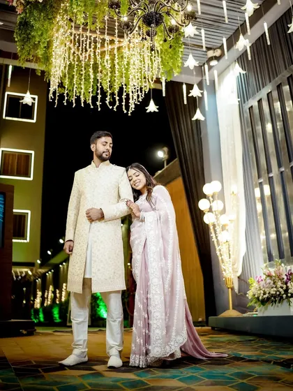 A full-length portrait of the couple at their reception, standing under a beautiful floral chandelier.