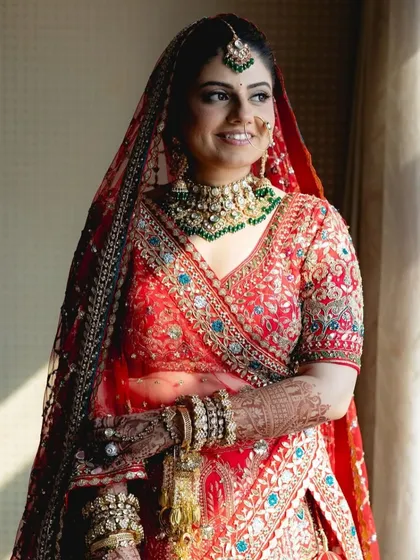 This bride looks serene and beautiful, framed by the window light. Her traditional red lehenga is perfectly accessorized with a green-accented kundan choker set, creating a picture of bridal grace.