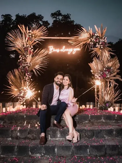 A wide shot of the entire proposal scene at night, with the floral arch, neon sign, and hundreds of candles creating a magical glow.