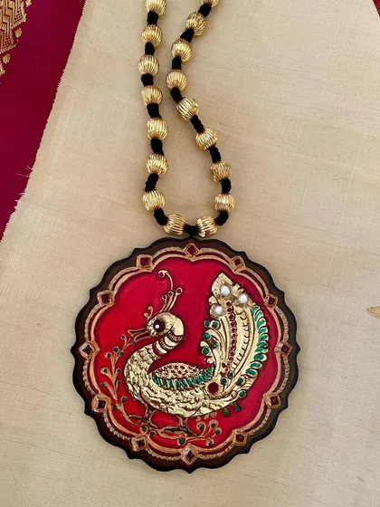 A majestic peacock, brought to life with real Tanjore gold foil against a rich red background. This wearable art piece is sealed with resin to protect its intricate gesso work and brilliant shine, making it a truly royal accessory.
