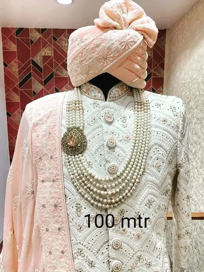 A beautiful pastel peach sherwani set displayed on a mannequin. This outfit features intricate white embroidery and is paired with a matching safa and a multi-layered pearl necklace, ideal for a modern groom.